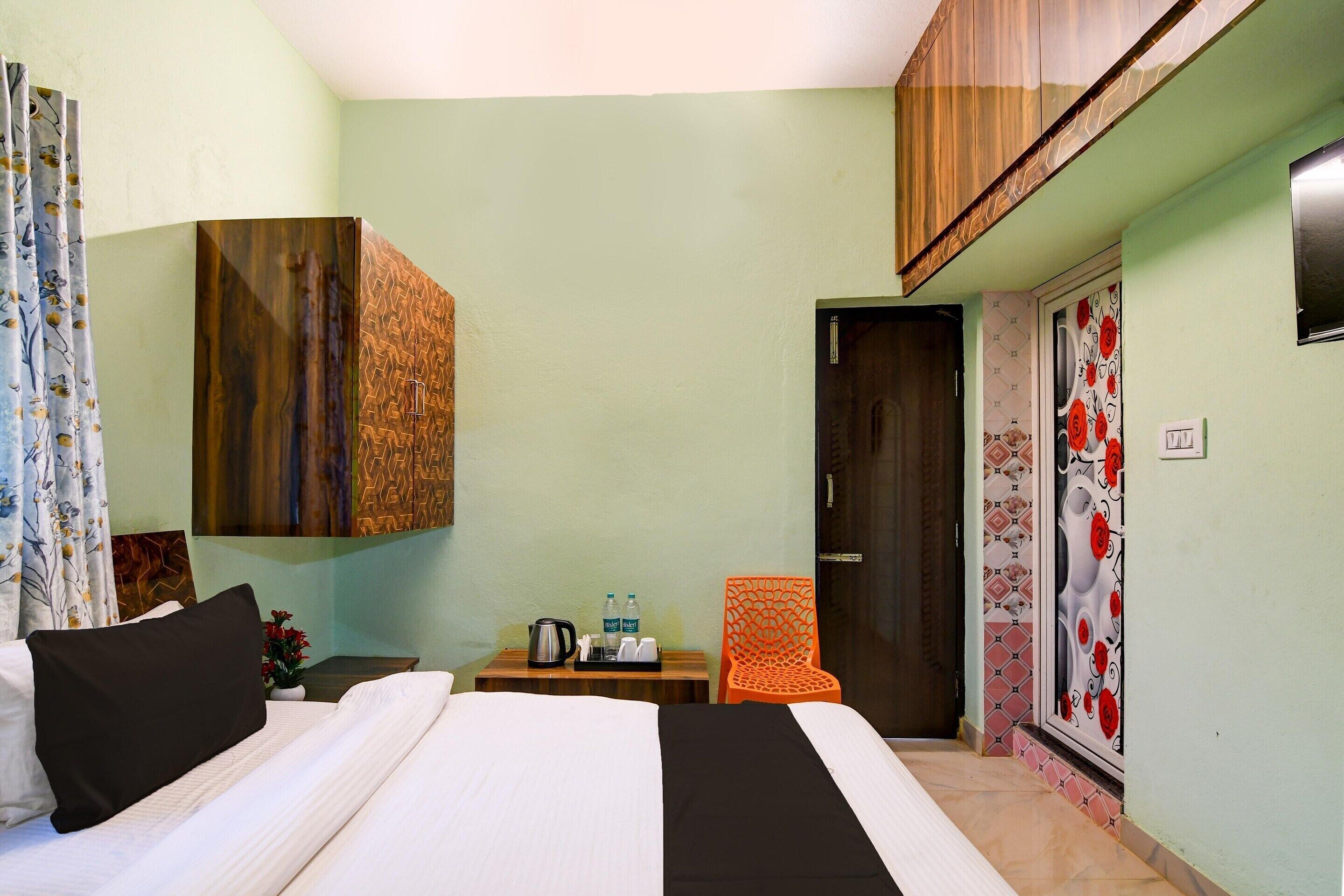 A hotel room with a bed, a small table with a kettle and water bottles, an orange chair, and a wardrobe with a geometric pattern.
