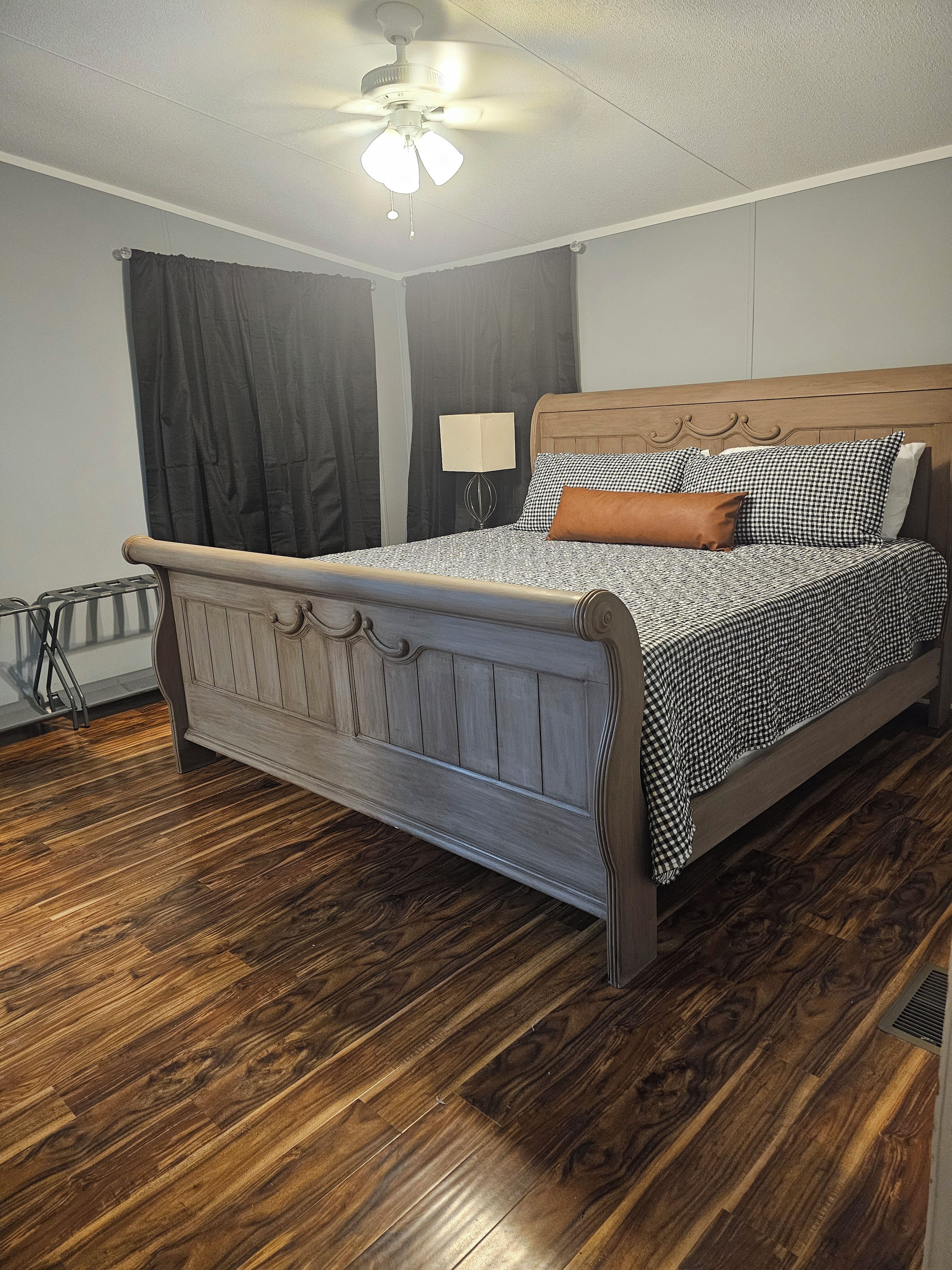 Un dormitorio con una cama de madera, un ventilador de techo, una mesita de noche con lámpara y cortinas oscuras.