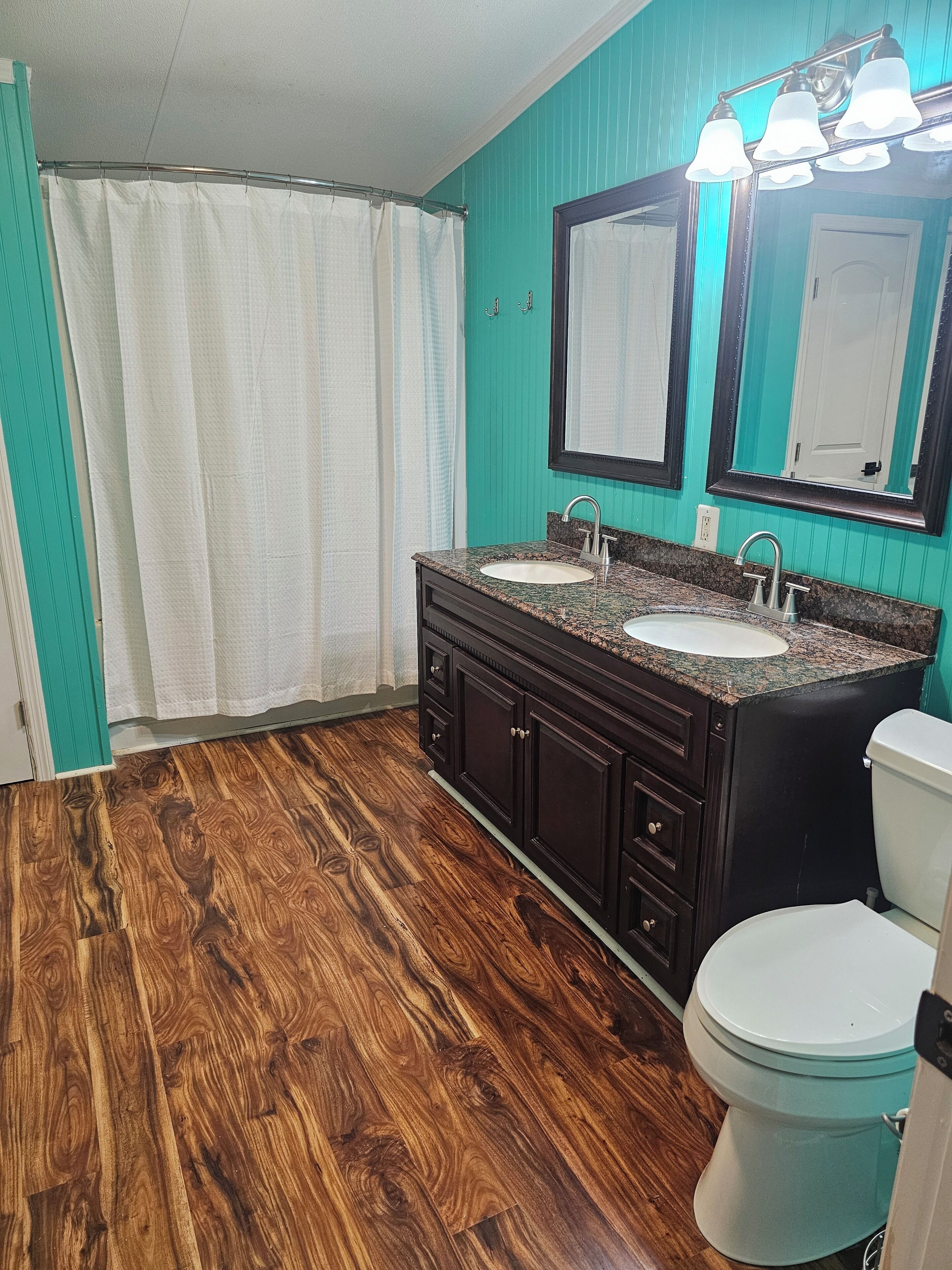 Un baño con piso de madera, dos lavamanos, un inodoro y cortina de ducha.
