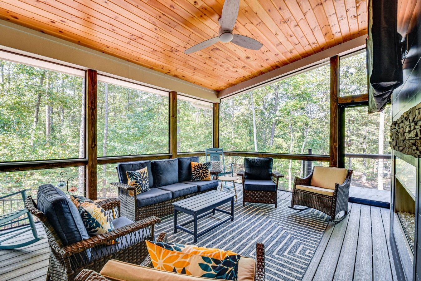 A covered outdoor seating area with a wooden ceiling, large windows, and a view of the forest.