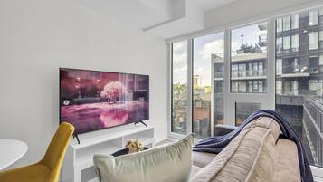 En moderne stue med et fladskærms-tv, en sofa, en gul stol og et stort vindue med udsigt til bygninger.