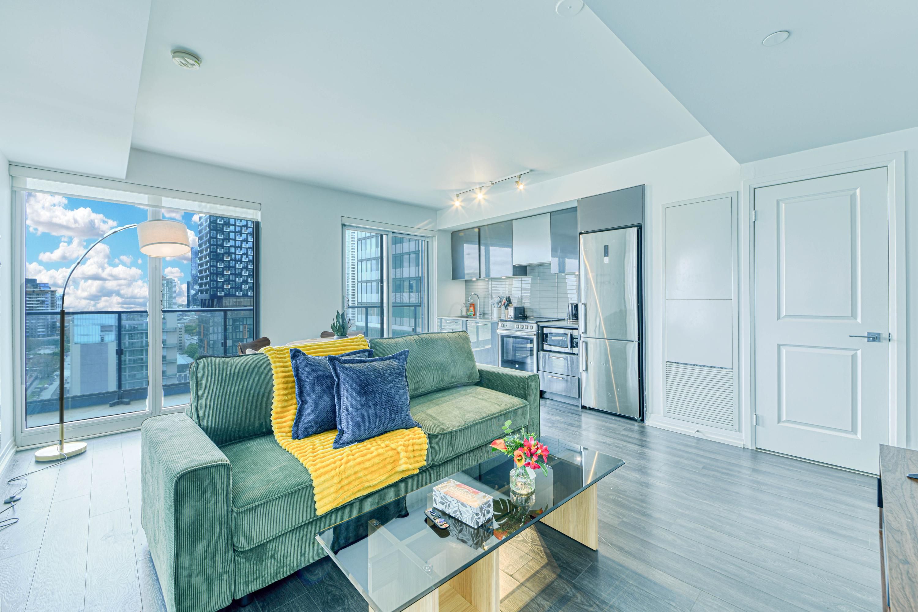 A modern apartment with a green sofa, a glass coffee table, and a view of the city through large windows.