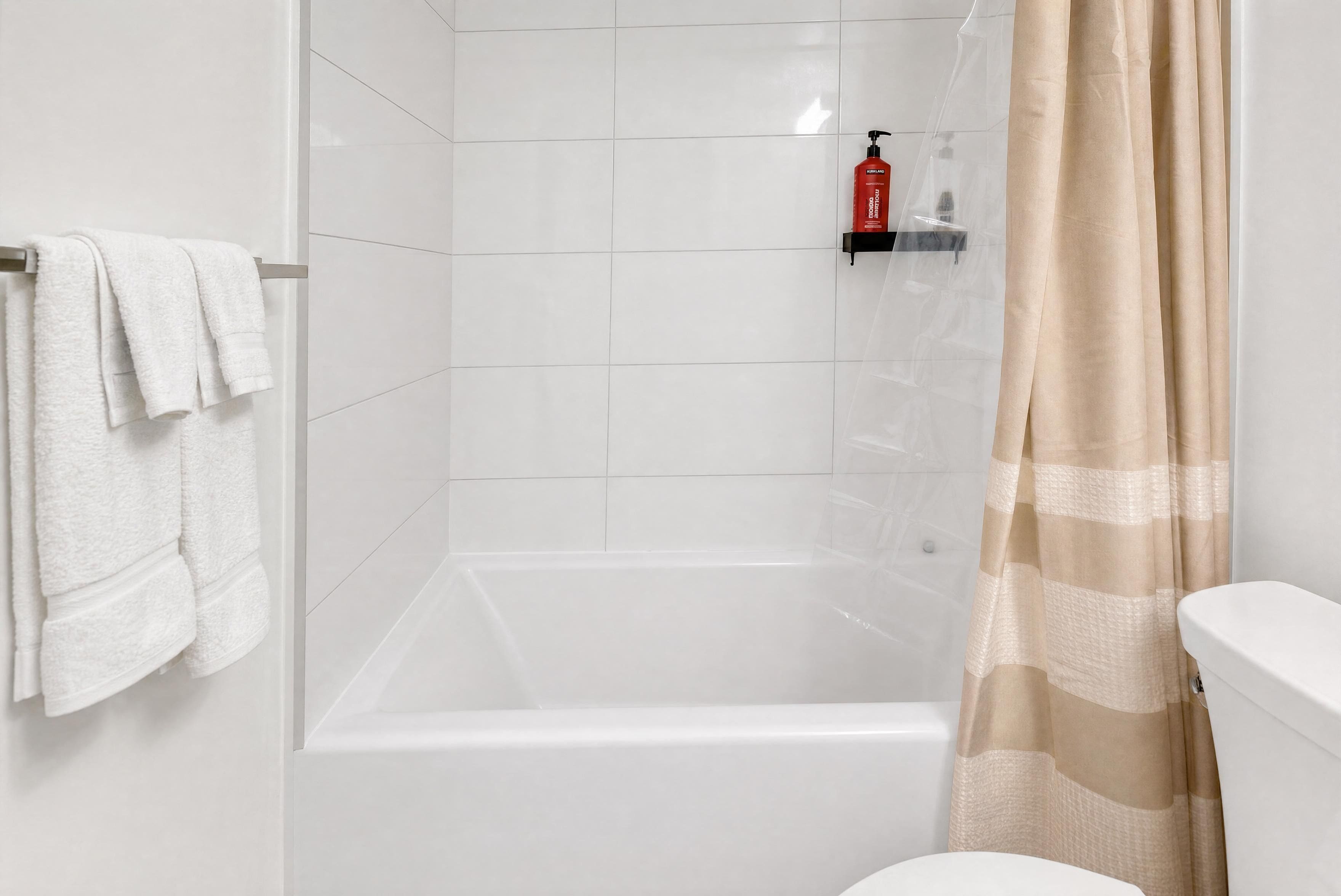 A shower area with a glass door, a white tiled wall, and a toilet with a beige shower curtain.