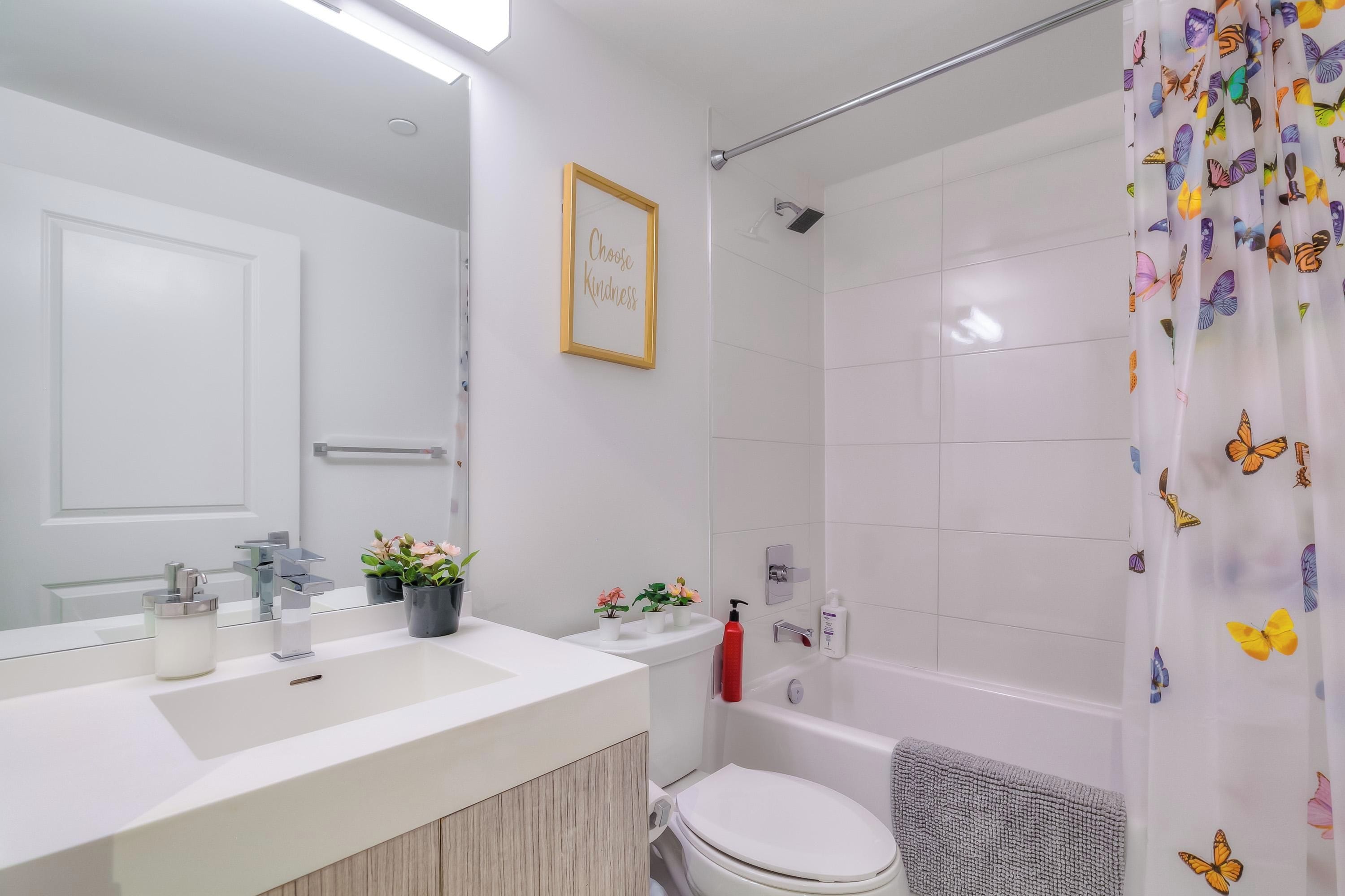 A modern bathroom with a white sink, mirror, and toilet. There is a shower curtain with a butterfly pattern, and a framed inspirational quote on the wall.