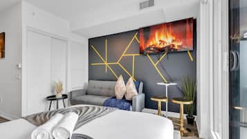 Una habitación de hotel moderna con una cama, un sofá gris, una mesita y un televisor de pared que muestra una chimenea.