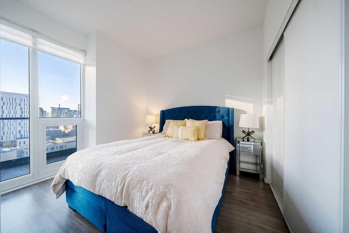 A modern bedroom with a large bed, two bedside tables with lamps, and a city view through large windows.