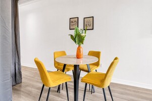 Een eetruimte met een ronde tafel, vier gele stoelen, een vaas met bloemen en twee ingekaderde foto's aan de muur.