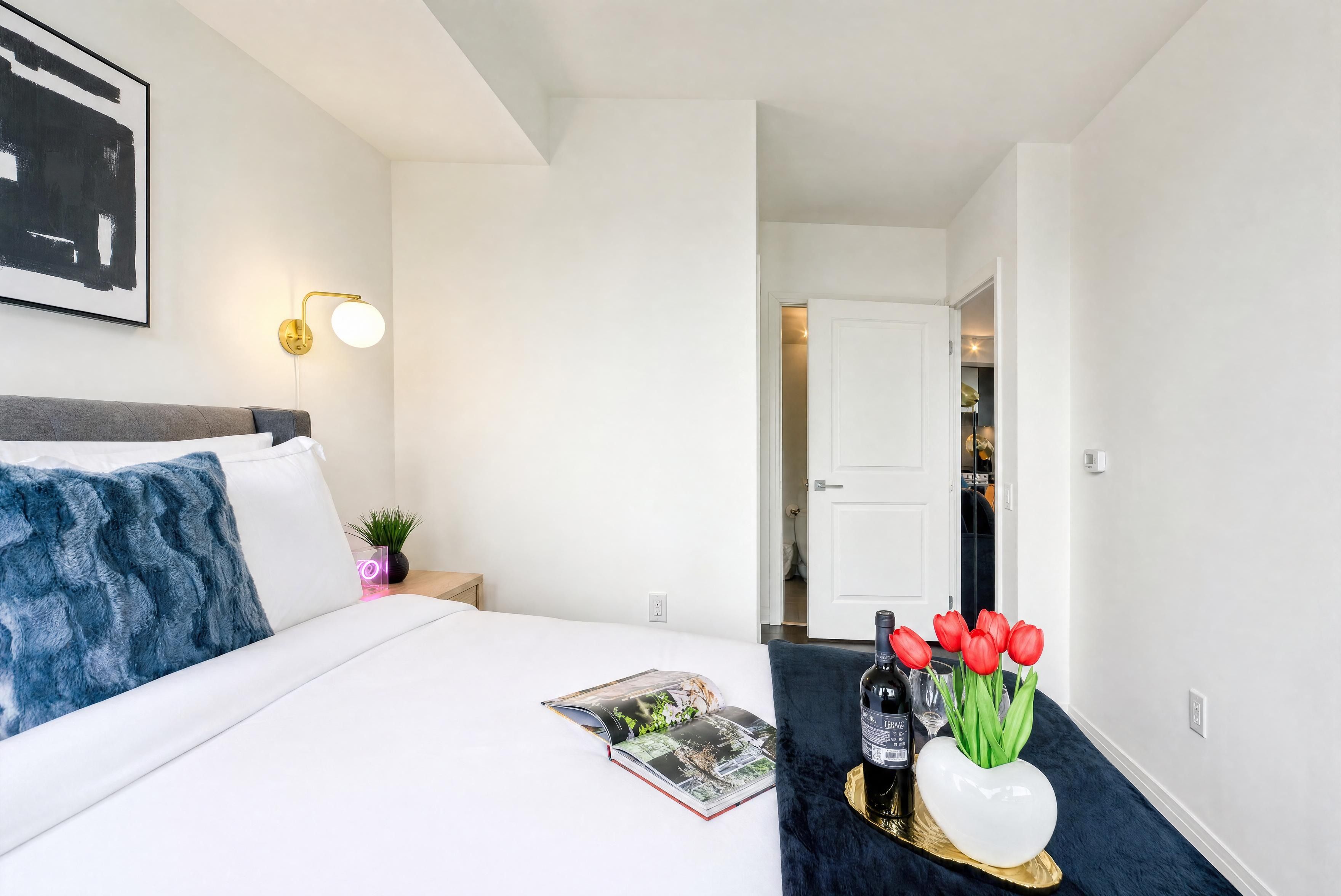 Et hotelværelse med en seng, et natbord med lampe, et magasin, en vase med røde blomster og en flaske vin.