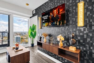 Een moderne woonkamer met een grote televisie waarop een filmplakat staat, een stadszicht door een groot raam en een decoratieve console tafel.