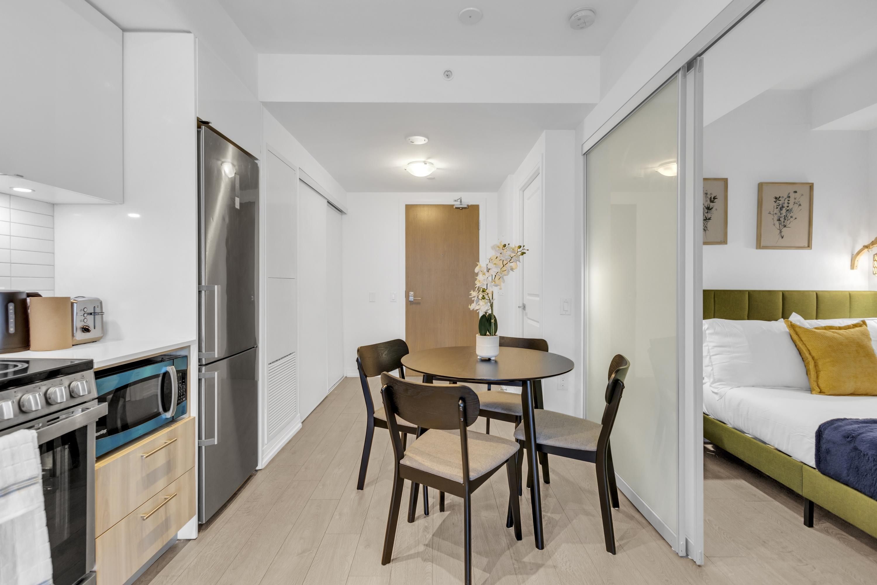 Une cuisine compacte dotée d’appareils électroménagers modernes, un coin repas avec une table ronde et des chaises, et un lit avec une tête de lit verte.