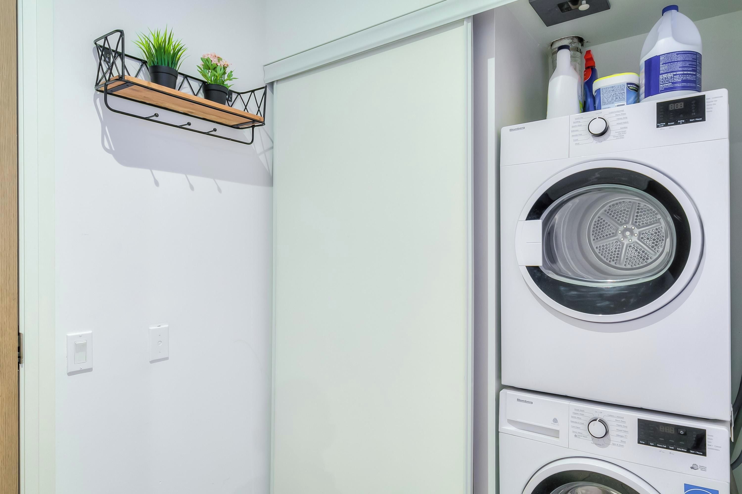 A laundry room with a stacked washer and dryer, a shelf with potted plants, and a water bottle on top.