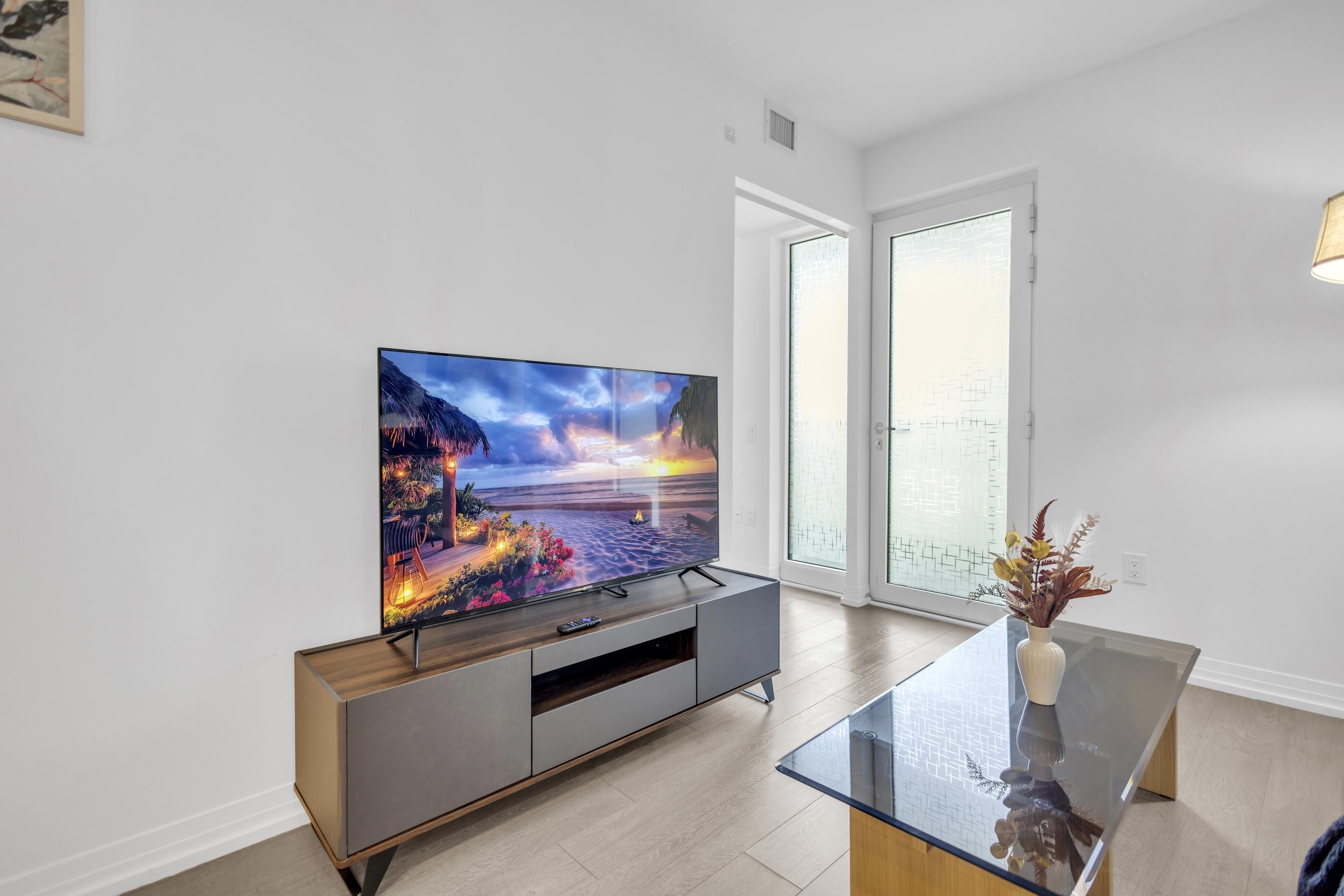 A modern living room with a flat-screen TV on a wooden cabinet, a glass coffee table, and a view of the outdoors through large glass doors.