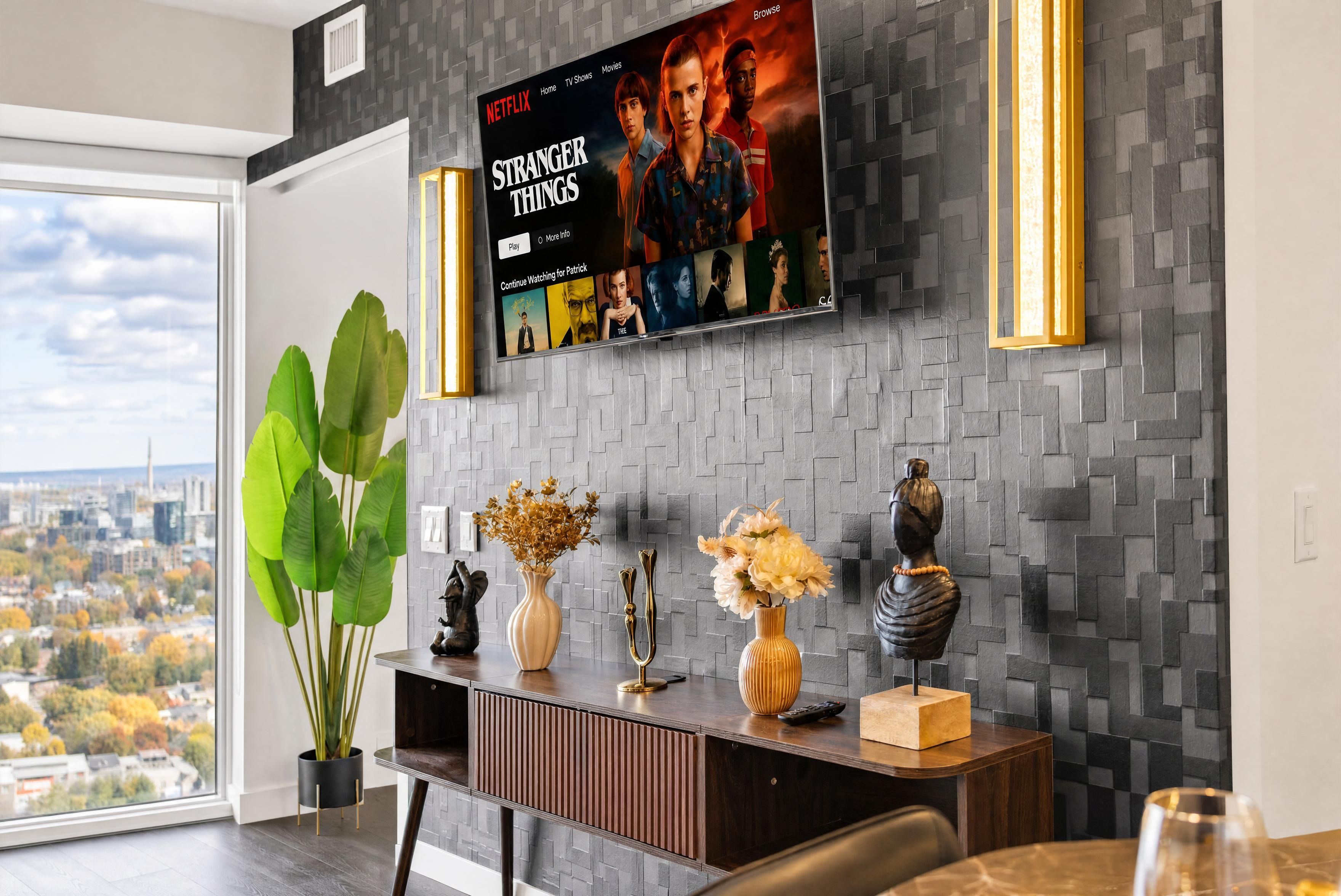 Una sala moderna con un televisor de pantalla plana grande montado en una pared texturizada, una mesa consola de madera con objetos decorativos y un ventanal con vistas a la ciudad.