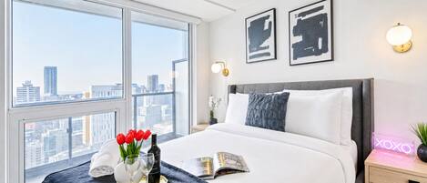 Une chambre d’hôtel avec une grande fenêtre, un lit recouvert de draps blancs, un plateau orné de fleurs rouges et deux œuvres d’art encadrées accrochées au mur.