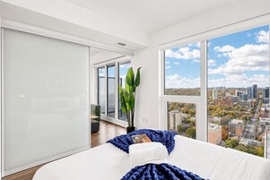 A modern bedroom with a large window, a bed, a potted plant, and a view of a cityscape.