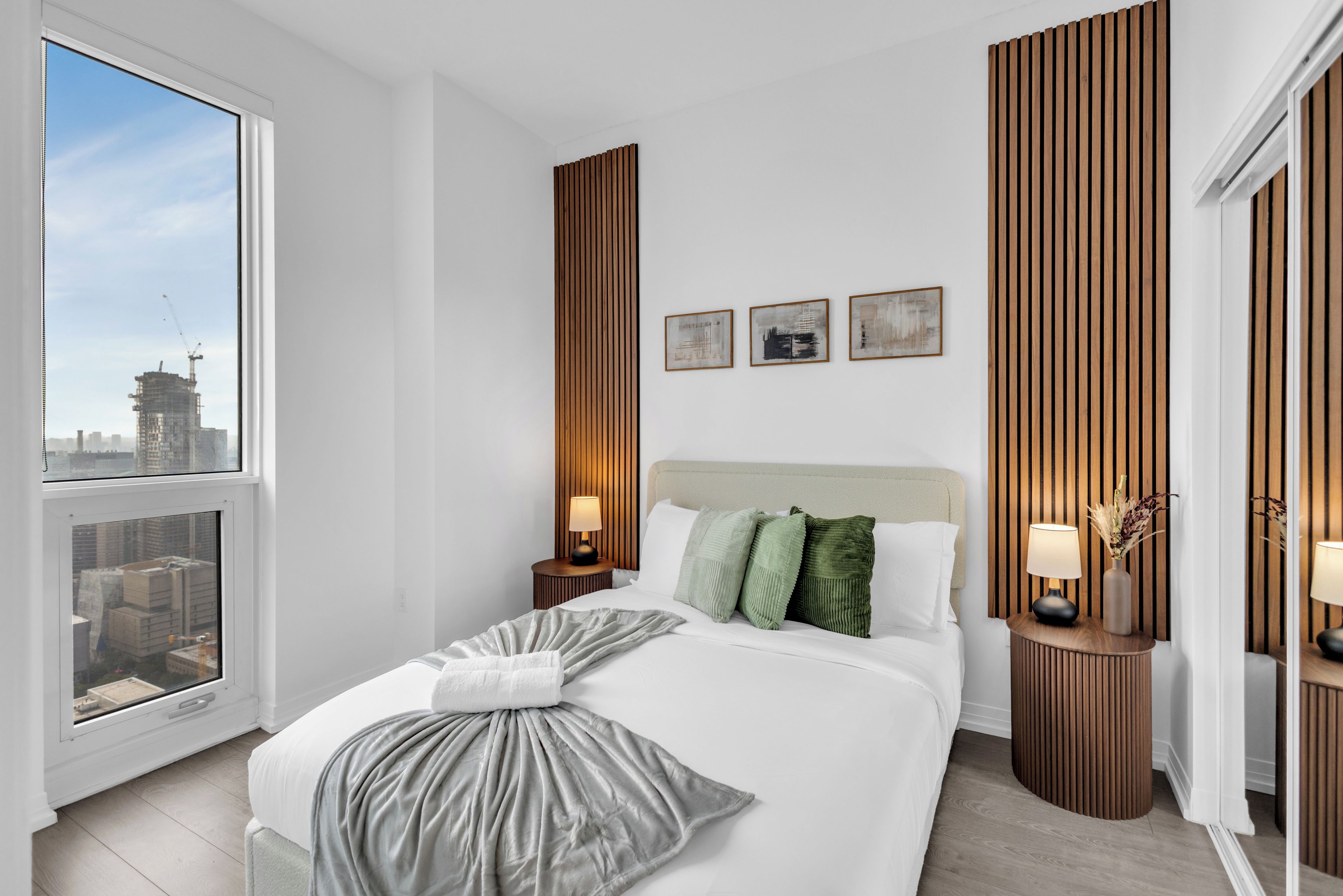 A modern hotel room with a large bed, wooden paneling, a city view, and three framed artworks on the wall.