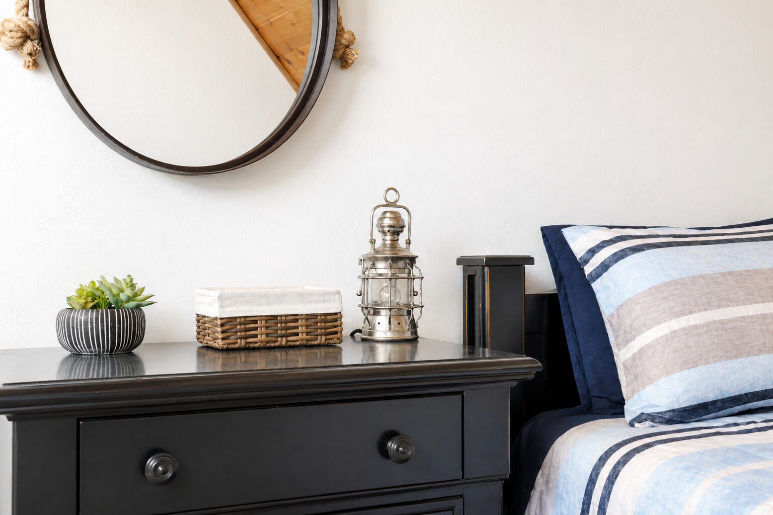 Un dormitorio con cama, mesita de noche con lámpara, espejo y una planta pequeña.