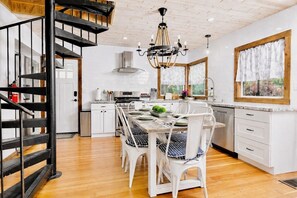 A modern kitchen with white cabinetry, a dining area with white chairs, and a black spiral staircase. - Lake Access Cabin, Walk to Trails + AC & Firepit (Lake Arrowhead)