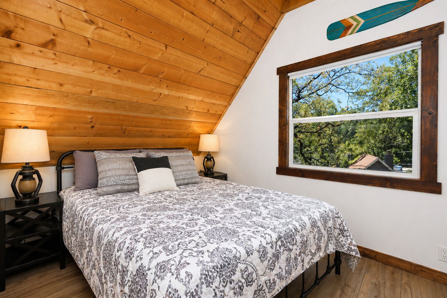Une chambre avec un plafond en bois, un lit recouvert d’une literie à motifs, deux lampes de chevet et une fenêtre donnant sur des arbres.