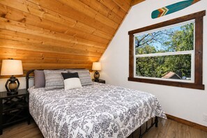 Ein Schlafzimmer mit Holzboden, einem Bett mit gemusterten Betttextilien, zwei Nachttischlampen und einem Fenster mit Blick auf Bäume.