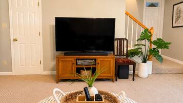 Una sala de estar con un televisor sobre un mueble de madera, una planta en maceta y una cesta con controles remotos.