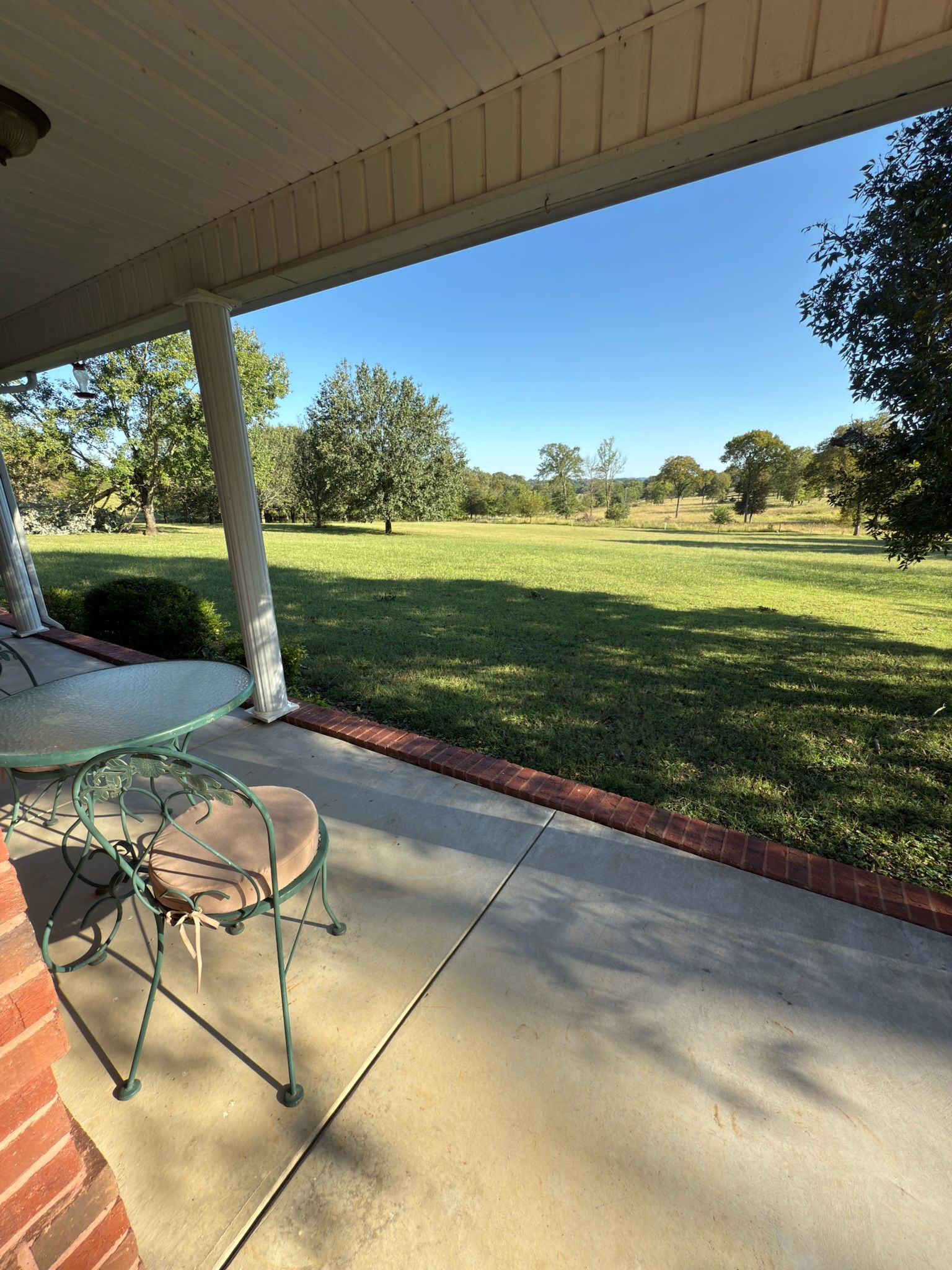 Un patio techado con piso de concreto, una silla de metal y vista a un campo cubierto de césped y árboles.
