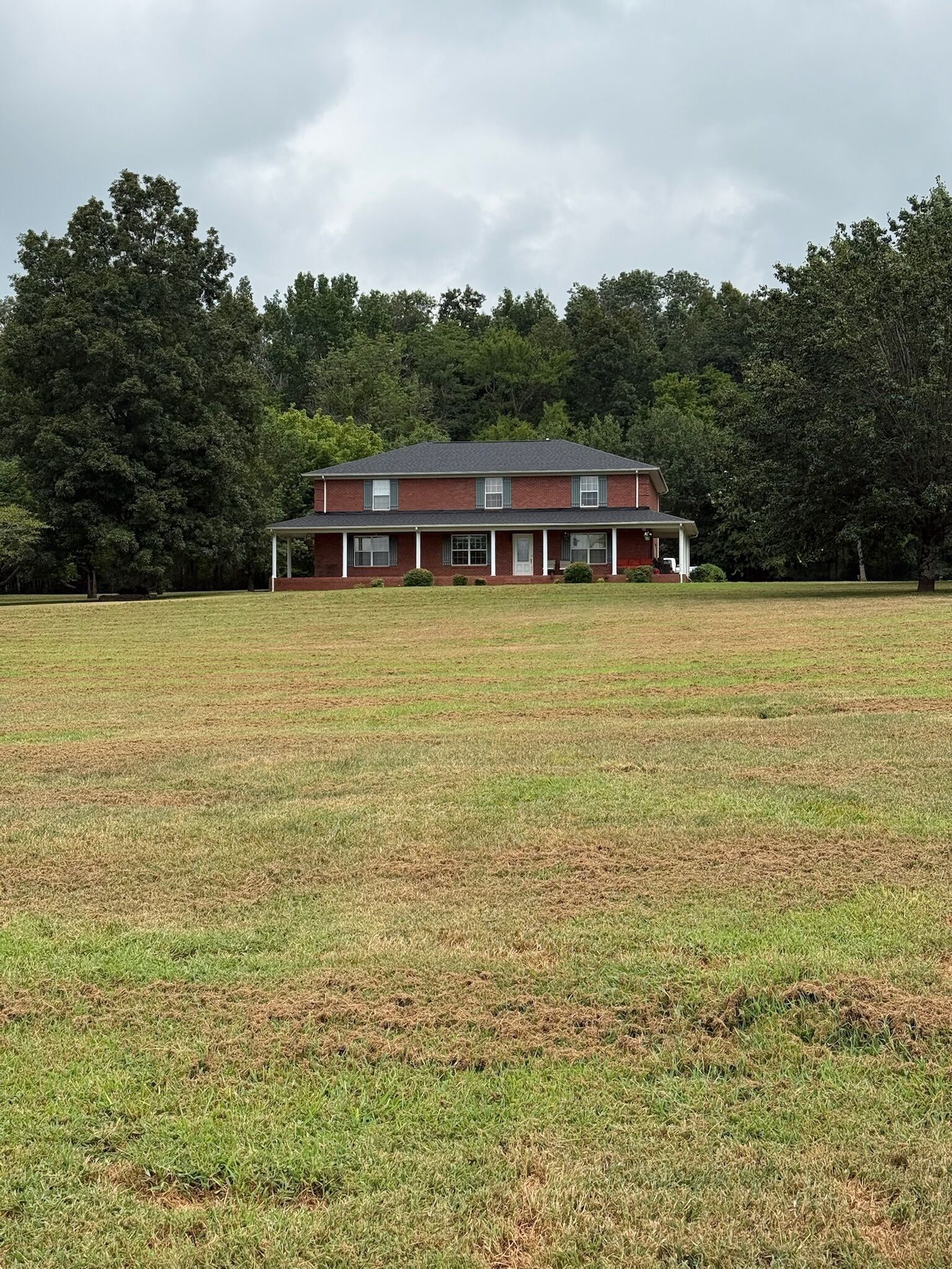 A single-story brick house with a front porch, surrounded by a large grassy field and dense trees in the background.