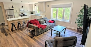 A modern kitchen with white cabinets, a stainless steel refrigerator, and a dining area with a gray sofa and red cushions.