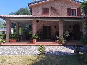 A two-story stone house with a covered porch, potted plants, and a stone-paved entrance.