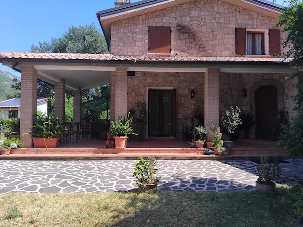 A two-story stone house with a covered porch, potted plants, and a stone-paved entrance.