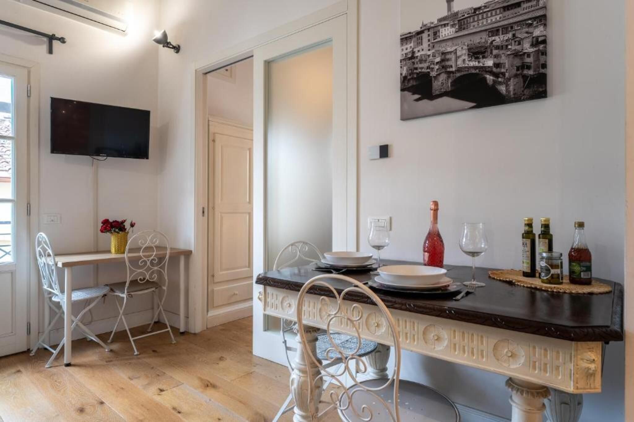 A dining area with a table set for two, a television mounted on the wall, and a framed picture above the table.