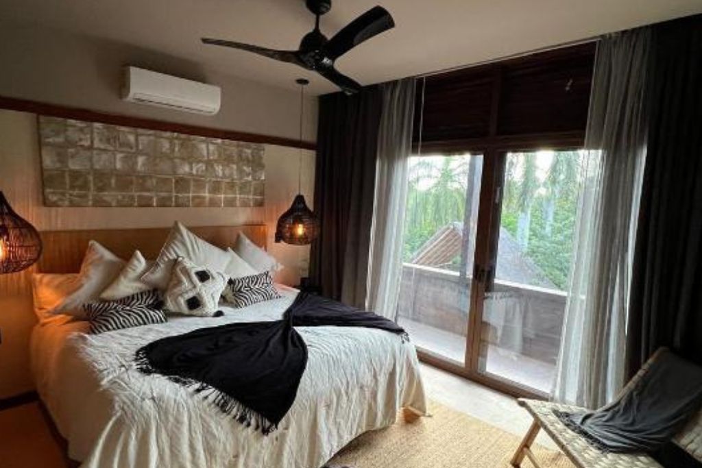 A bedroom with a bed, a ceiling fan, a window with curtains, and a view of greenery outside.