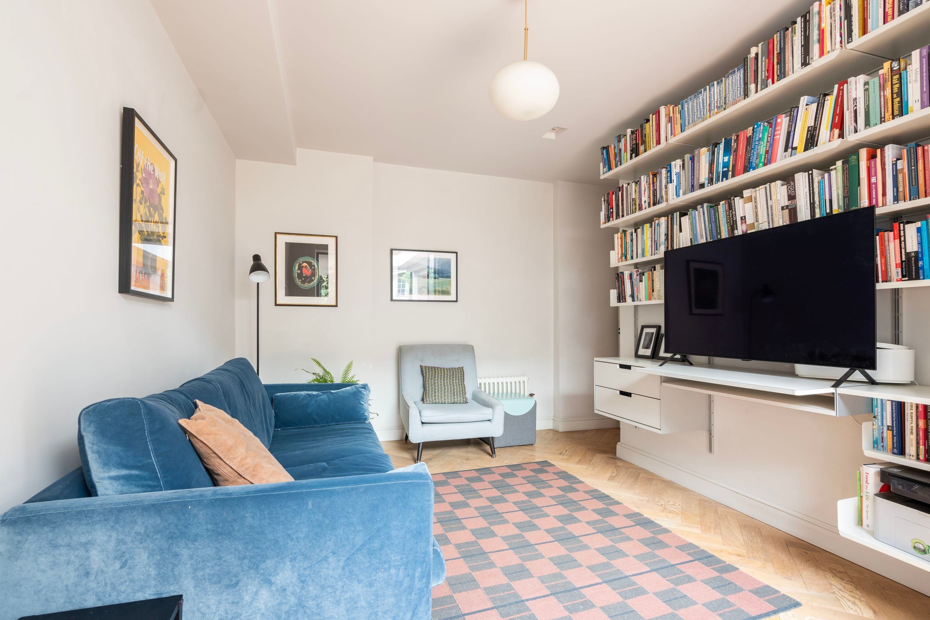 Een moderne woonkamer met een blauwe bank, een boekenplank vol boeken, een flatscreen televisie en een kamerkleed met een patroon.