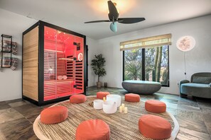 Ein modernes Wohnzimmer mit einem großen Glasvitrine, einem Deckenventilator, einem großen Fenster mit Blick auf Kakteen und mehreren runden, orangefarbenen Hocker.