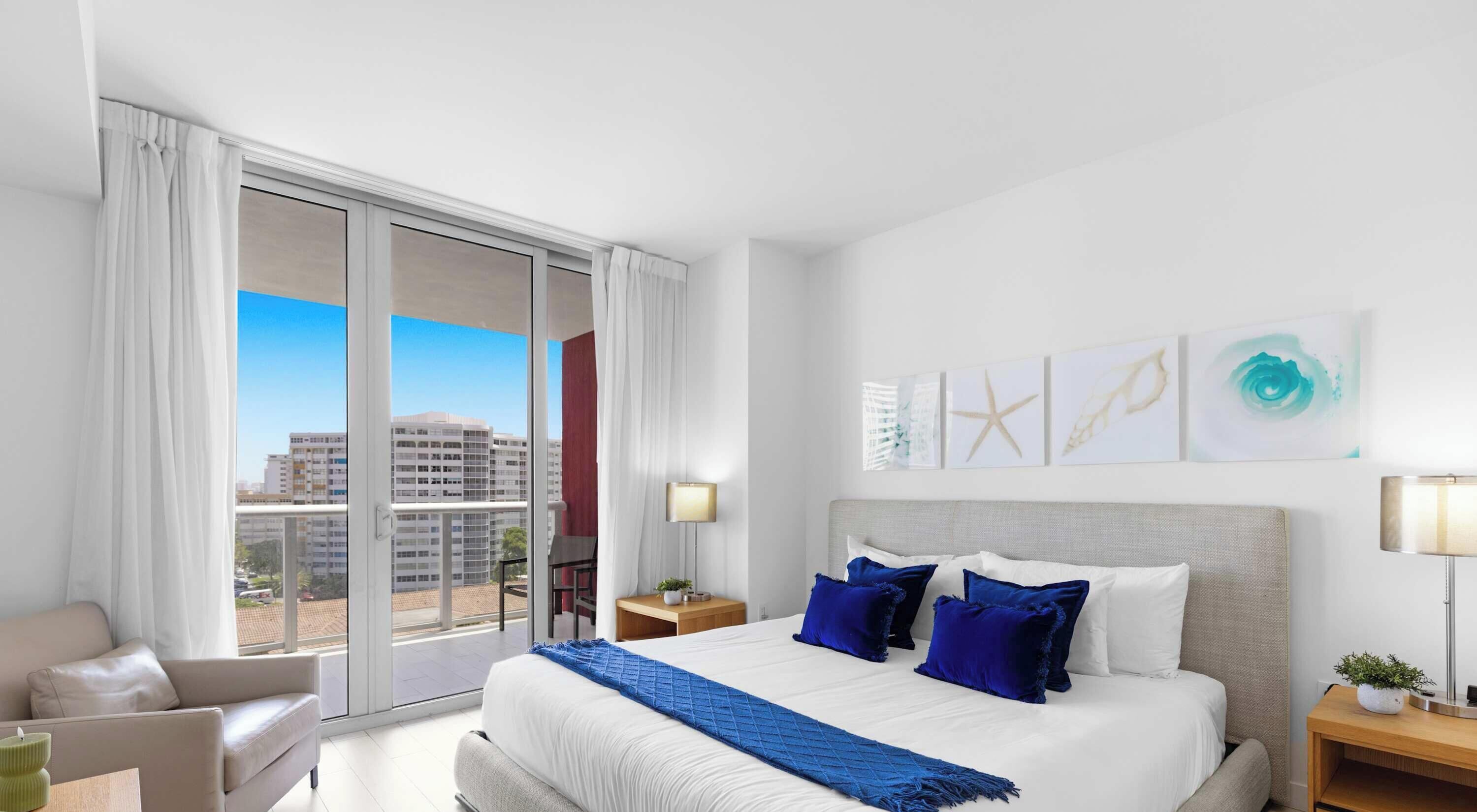 A hotel room with a large bed, a sofa, a view of buildings through the window, and artwork on the wall.