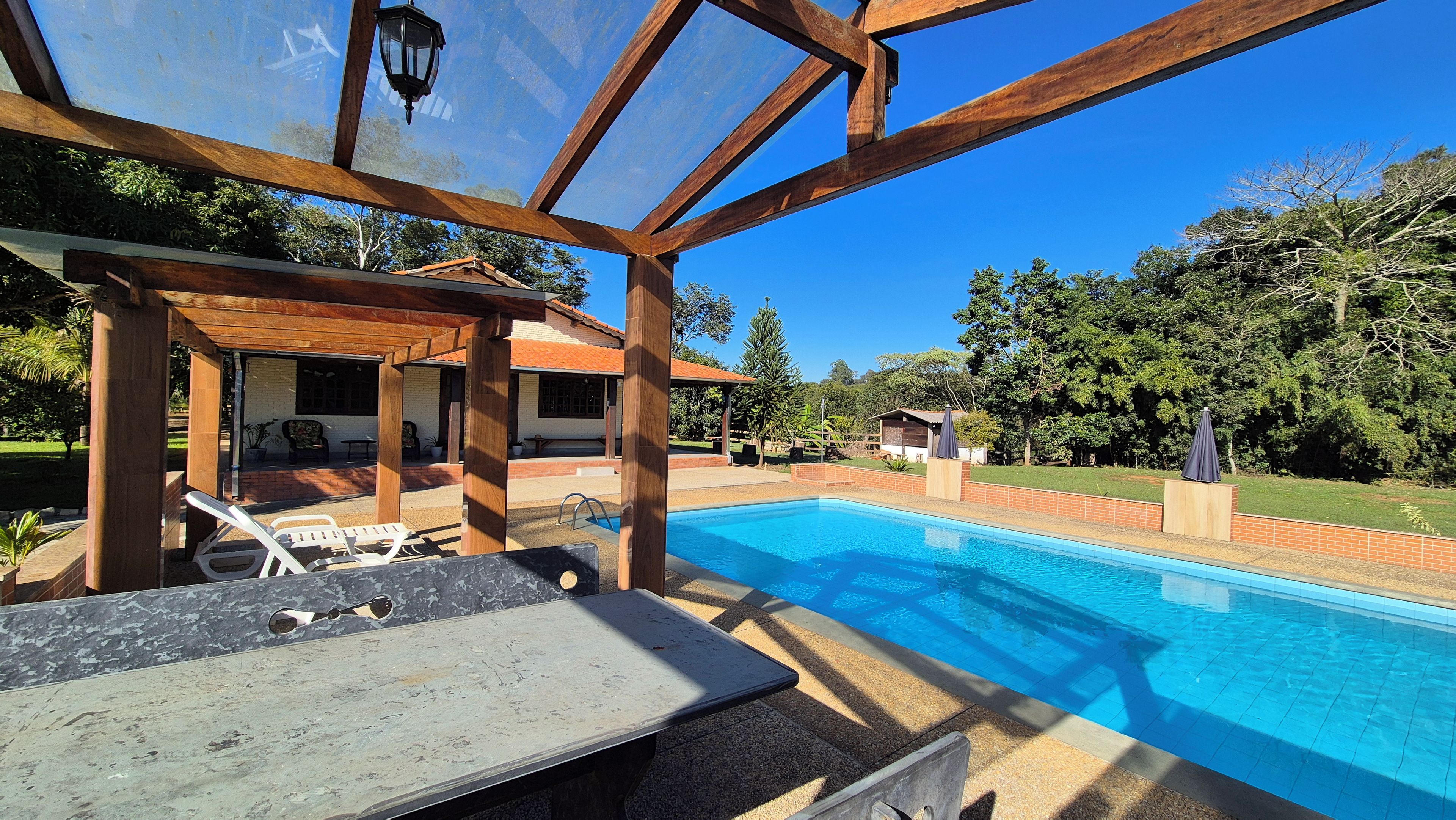Área de piscina com pergolado, assentos externos e céu azul límpido.
