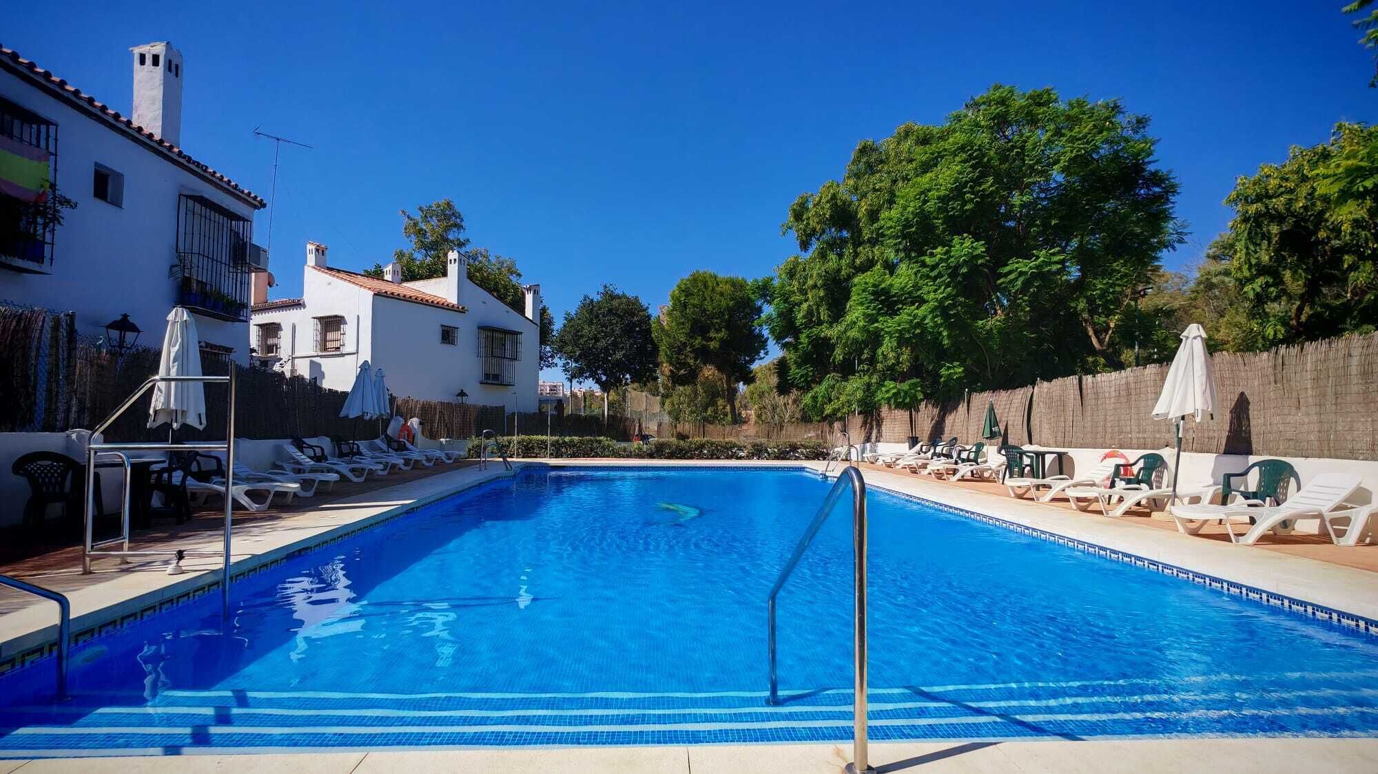 A swimming pool with lounge chairs and umbrellas, surrounded by a white building with a tiled roof.