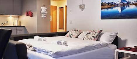 A hotel room with a bed, a desk, a chair, a small table, a painting of a mountain, and a heart-shaped decoration on the wall.