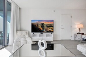 A modern living room with a flat-screen TV mounted on a white wall, a glass coffee table, and a white sofa. - Cozy and Restful 1- BR APT Steps from the Ocean (Hallandale Beach)