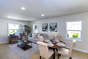 Un salon moderne comprenant un ensemble de canapés, une table basse et une table à manger pour quatre personnes. Deux fenêtres laissent entrer la lumière naturelle et offrent une vue sur l’extérieur.
