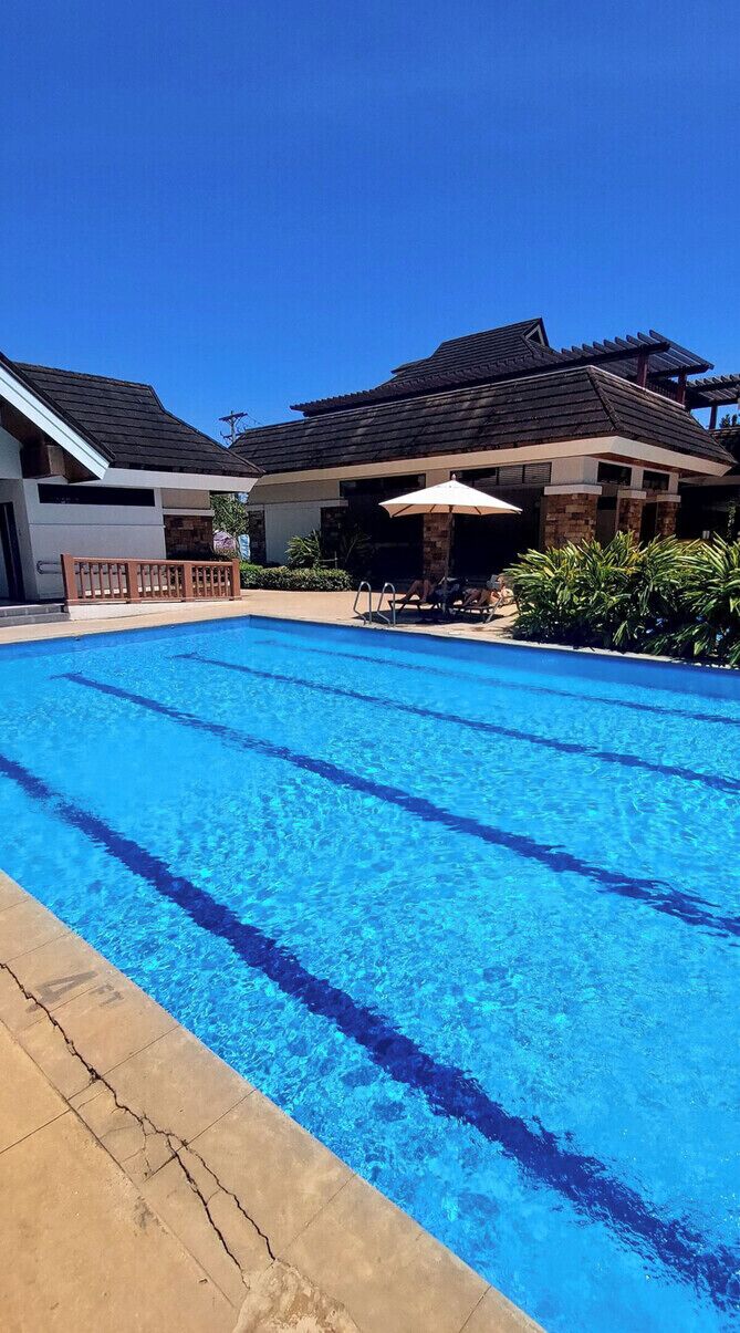 A swimming pool with clear blue water, surrounded by lounge chairs and umbrellas, with buildings in the background.