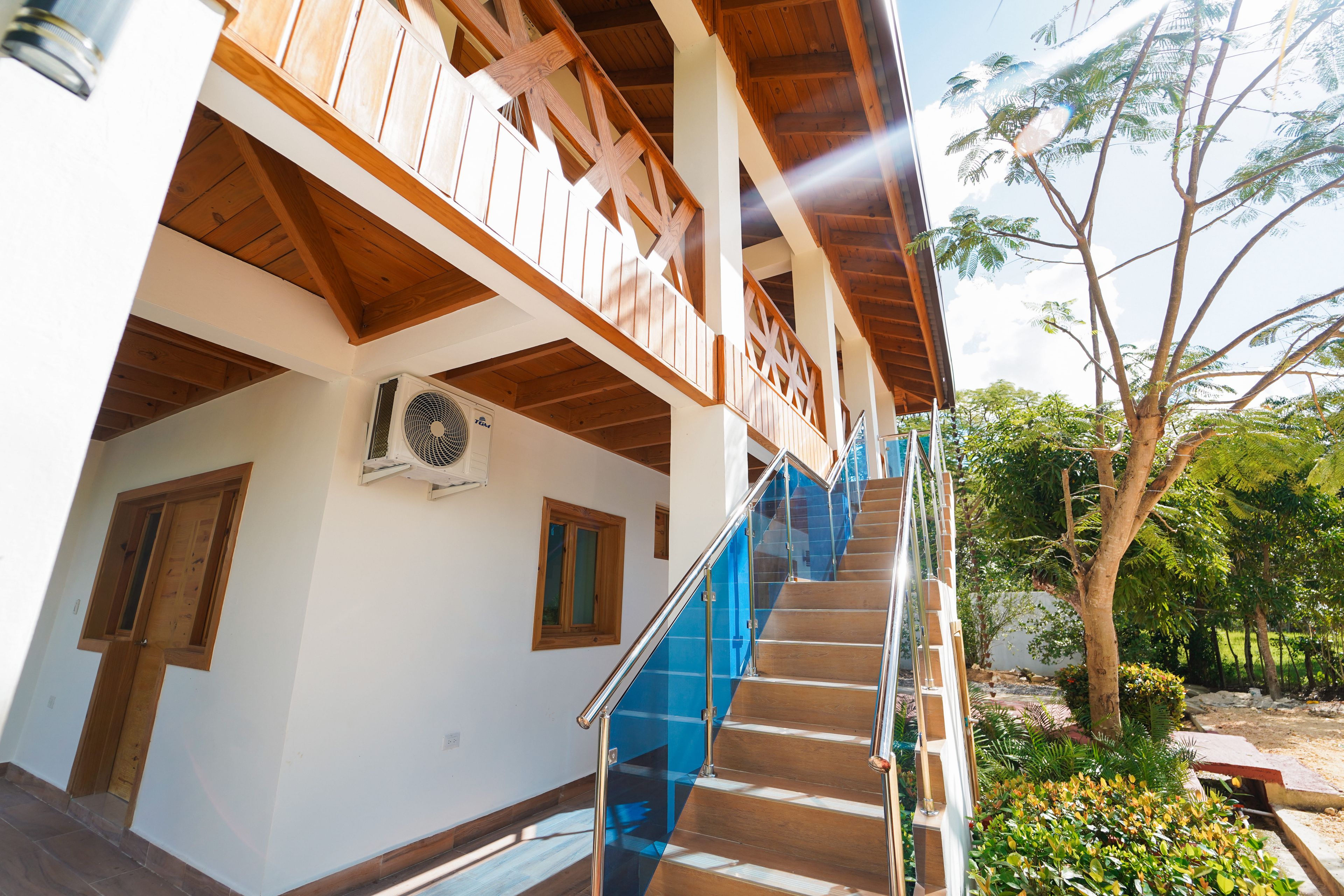 Un edificio de varios niveles con balcones de madera, una escalera con barandillas de vidrio azul y vistas a árboles y vegetación.