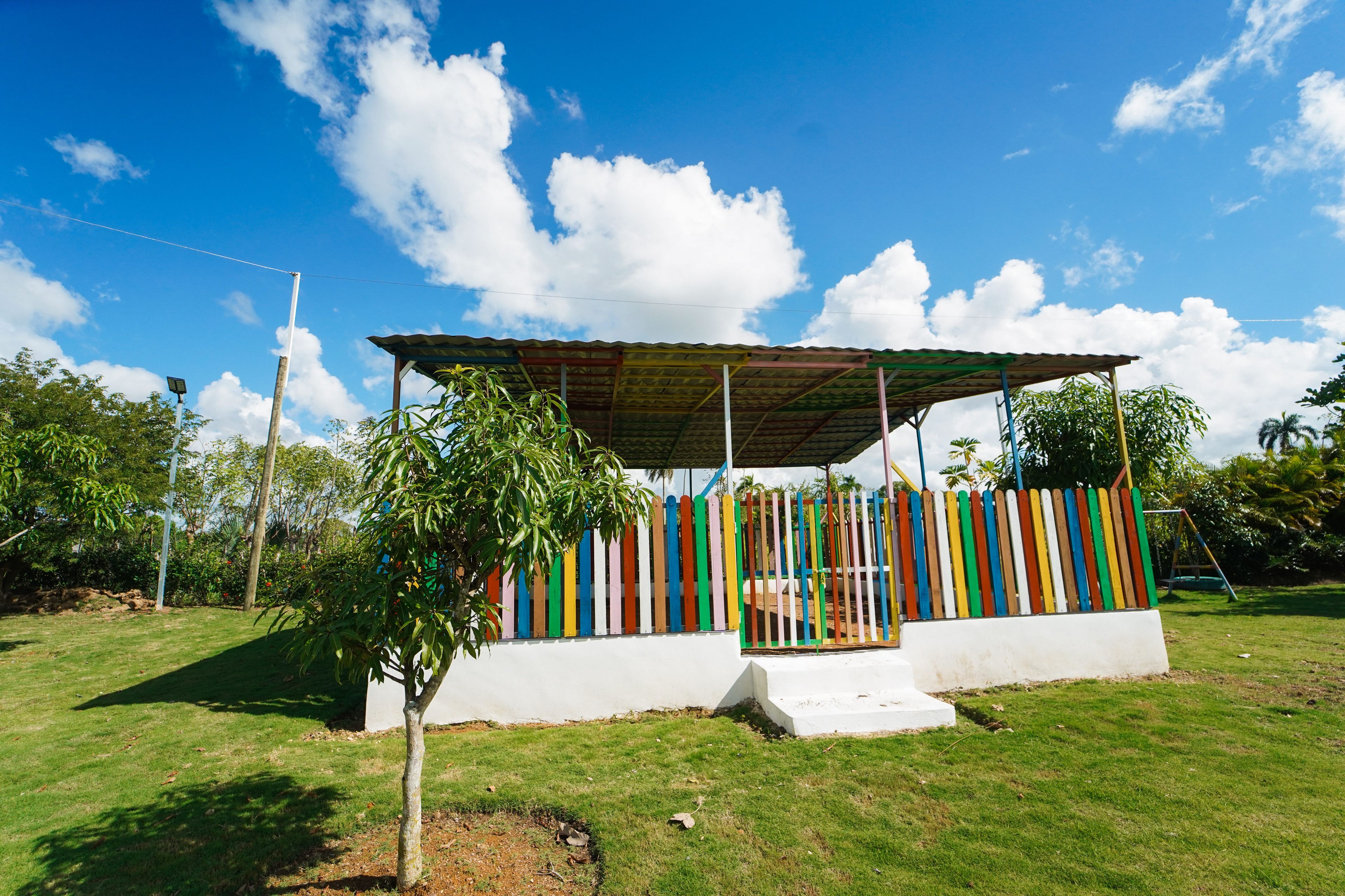 A small, open-air structure with a colorful fence and a roof, surrounded by grass and trees.