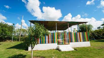 A small, open-air structure with a colorful fence and a roof, surrounded by grass and trees.