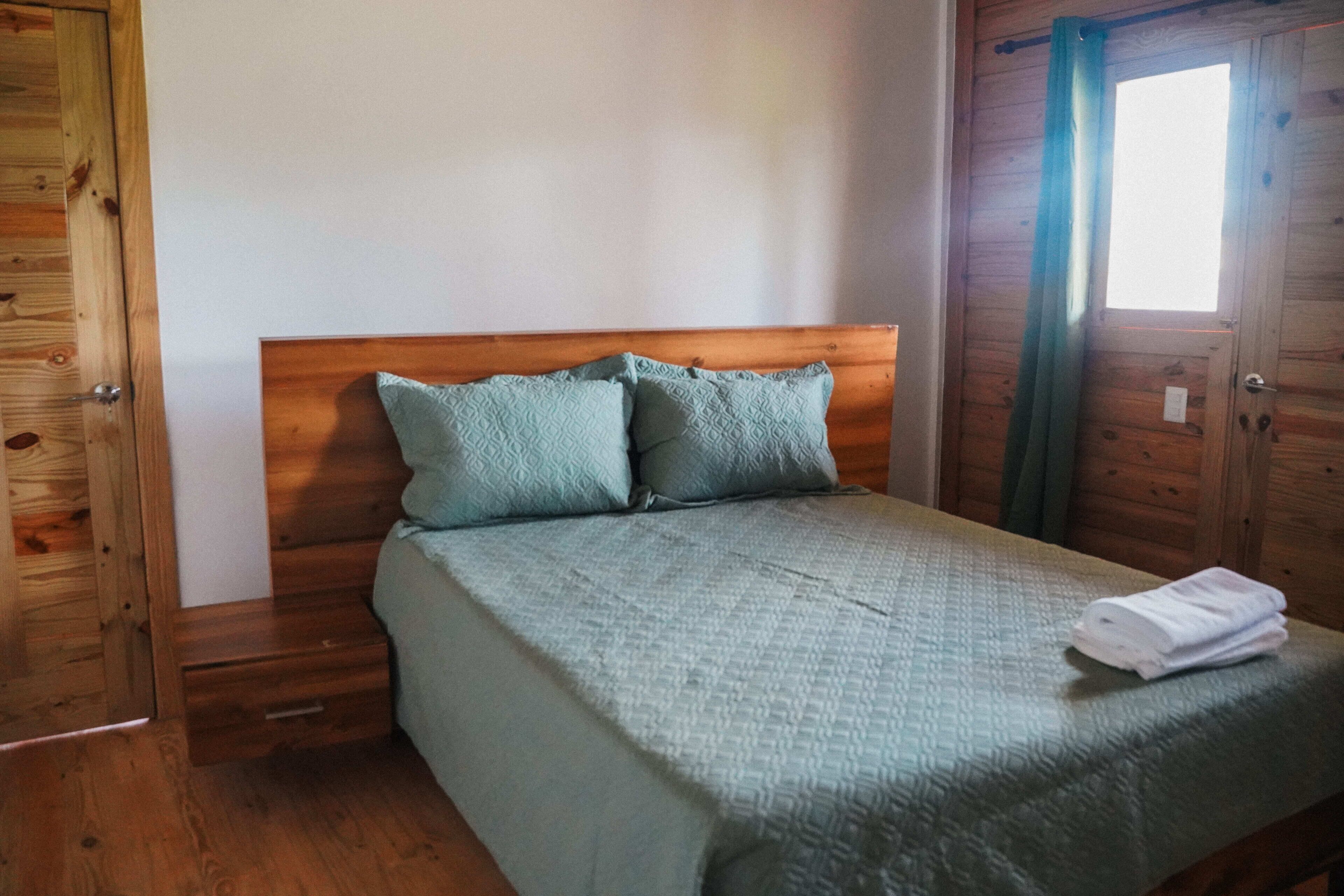 A wooden-framed room with a bed, two pillows, a folded towel, and a window with curtains.