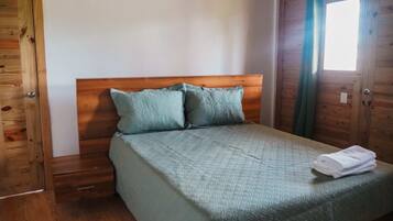 A wooden-framed room with a bed, two pillows, a folded towel, and a window with curtains.