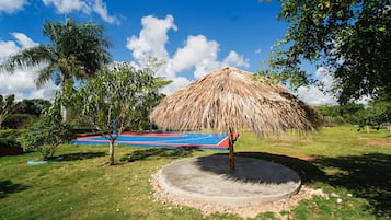 A thatched-roof structure with a blue and red floor, surrounded by grass and trees.