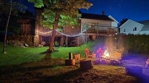 A backyard with a fire pit, a wooden deck, and a house with string lights.
