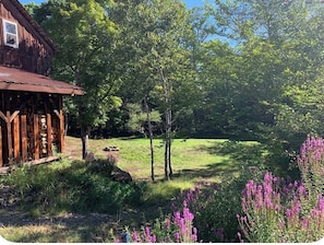 A rustic wooden cabin with a porch, surrounded by lush greenery and flowering plants.