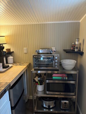 A compact kitchen with a built-in oven, a coffee maker, a kettle, and a slow cooker on a shelf.